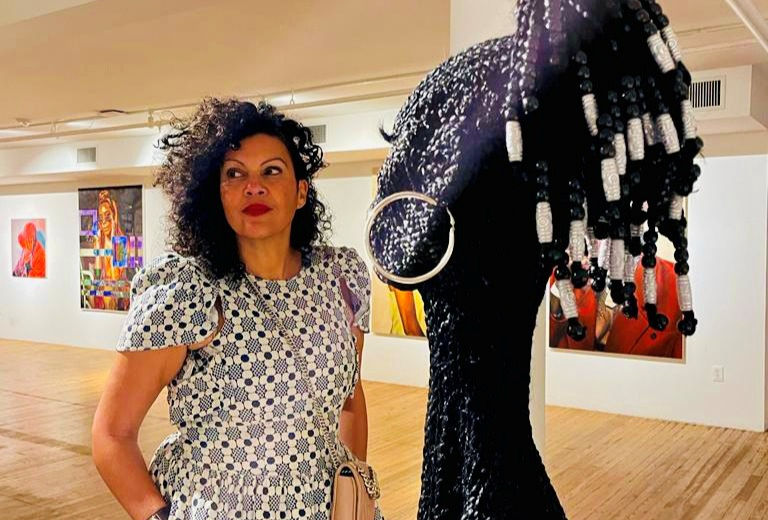Woman in patterned dress stands in an art gallery beside a large black beaded sculpture, looking thoughtful.
