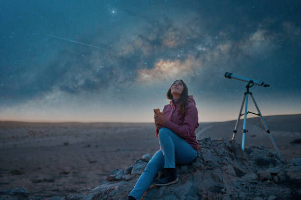 Woman in pink hoodie stargazes with telescope on rocky terrain; night sky and Milky Way visible, evoking a serene, contemplative mood.