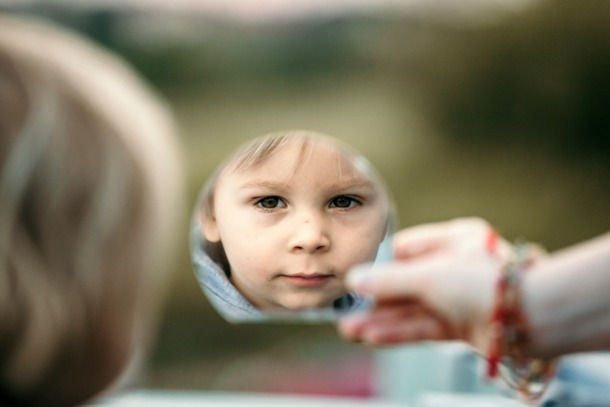 Child’s face reflected in a small handheld mirror, with a hand holding it outdoors against a blurred green background.