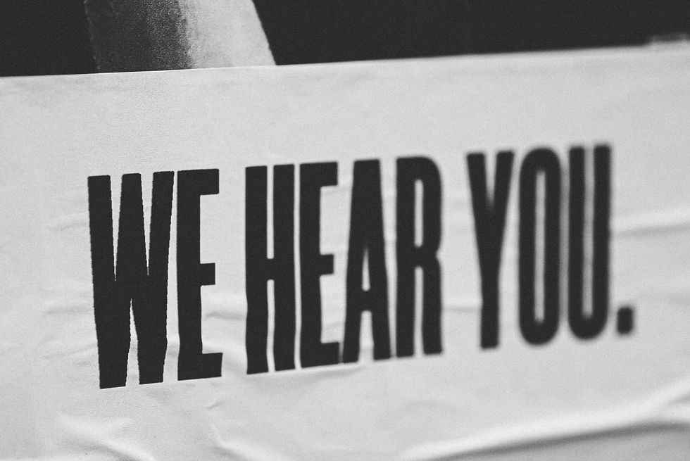Black and white image of a sign with bold black text "WE HEAR YOU." on a wrinkled white background. Mood is attentive and supportive.