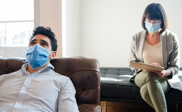 A masked man sits on a couch while a masked woman with a clipboard sits across from him.