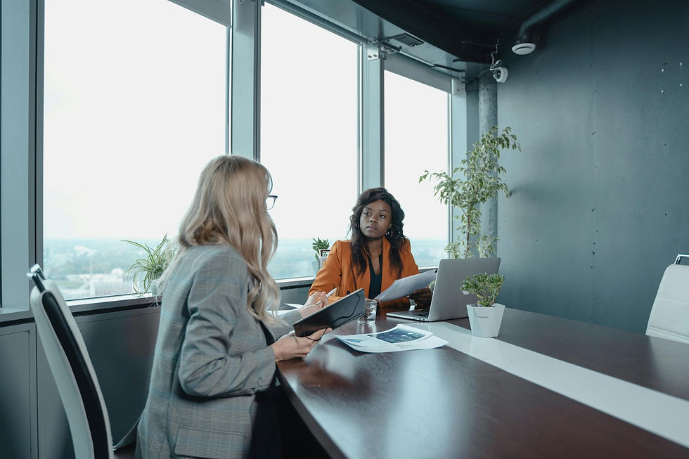 Two women in a modern office setting discuss papers. One in a gray blazer and holding a tablet, the other in an orange blazer, with a laptop.