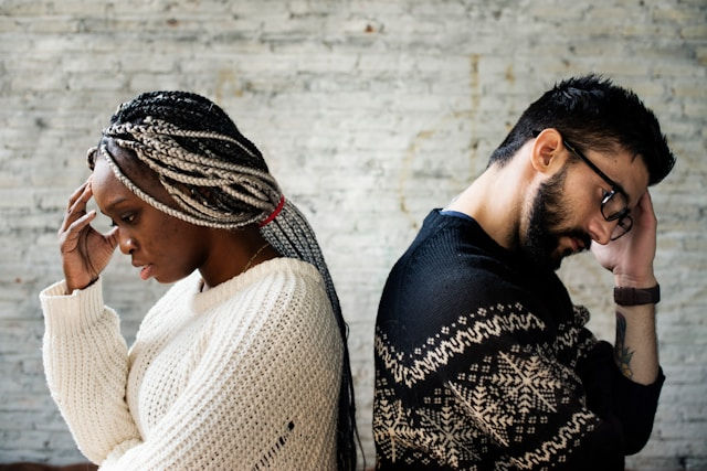 Two people stand back-to-back, looking pensive. Both touch their heads. They wear patterned sweaters against a white brick wall.