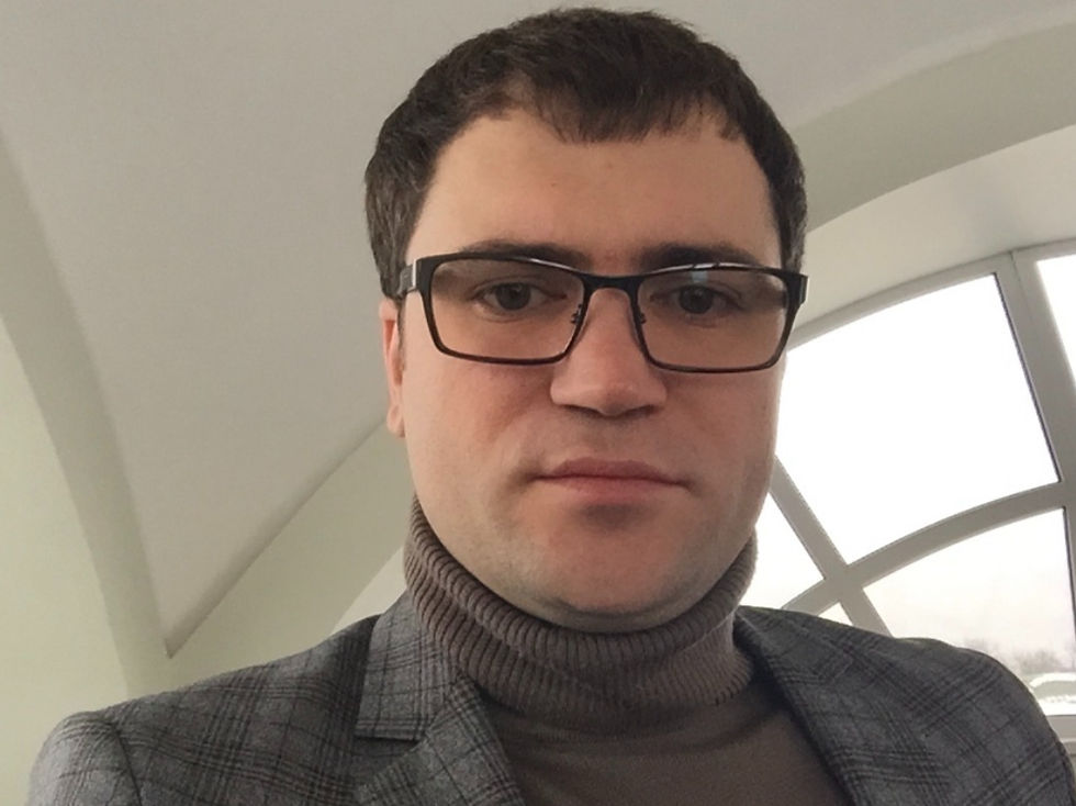 Close-up of a man in black glasses, gray plaid blazer, and turtleneck, indoors by a bright window, looking neutral.