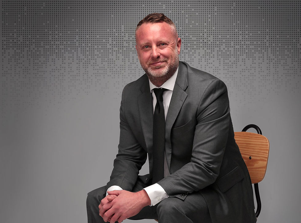 Christopher A. Smith in a gray suit and black tie sitting on a wooden chair, smiling slightly. Gray background with a subtle pixel pattern.