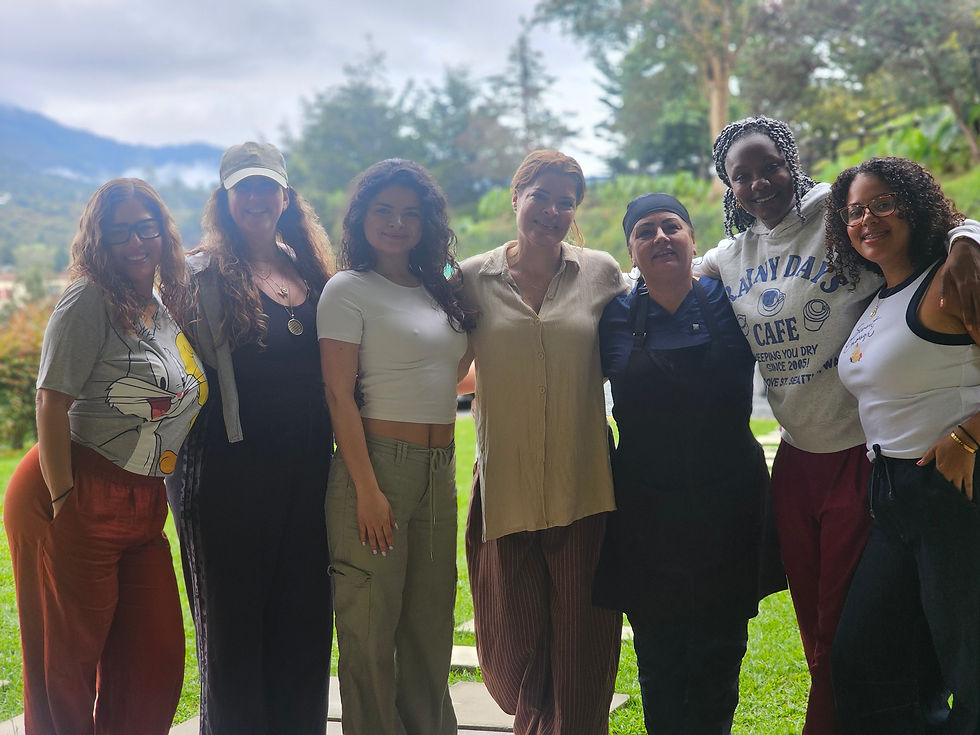 Seven people smiling, standing arm-in-arm outside with mountains and greenery in the background. Casual attire, relaxed and joyful mood.