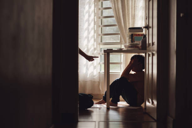 A little Asian boy with a sad and lonely expression hides in the shadows under a table at home, while being scolded by his mother