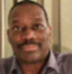 Man with a mustache and earrings, wearing a grey shirt, in a neutral interior setting. Calm expression, soft lighting.