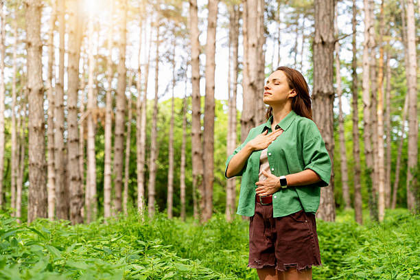 Woman in green shirt stands in a forest, eyes closed, hand on chest, looking calm among tall trees and green undergrowth.