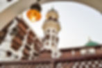 Ornate white and brown building with wooden lattice windows and a tall tower under a clear sky. An archway with glowing lights frames the view.