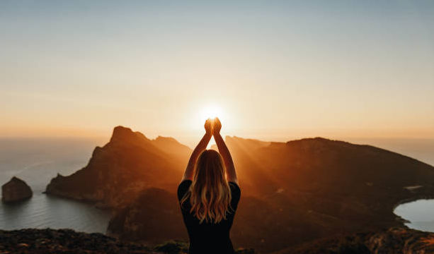 A woman stands on a mountain at sunset, holding her hands up as if cradling the sun.