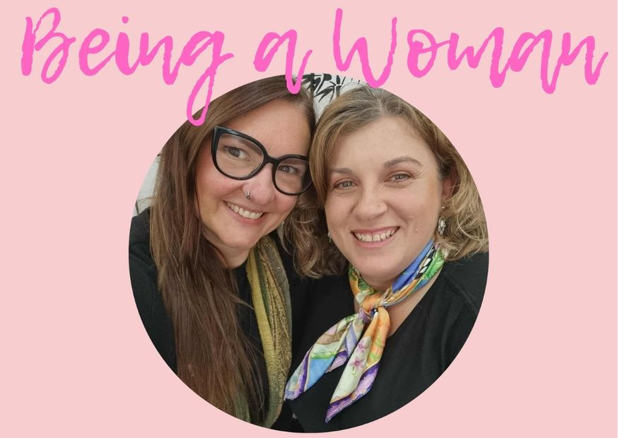 Two smiling women pose together on a pink background with "Being a Woman" text. One wears glasses; the other has a colorful scarf.