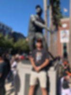 Man in Giants attire poses by a baseball statue in front of Oracle Park. Crowd and palm trees in background, sunny day, energetic vibe.
