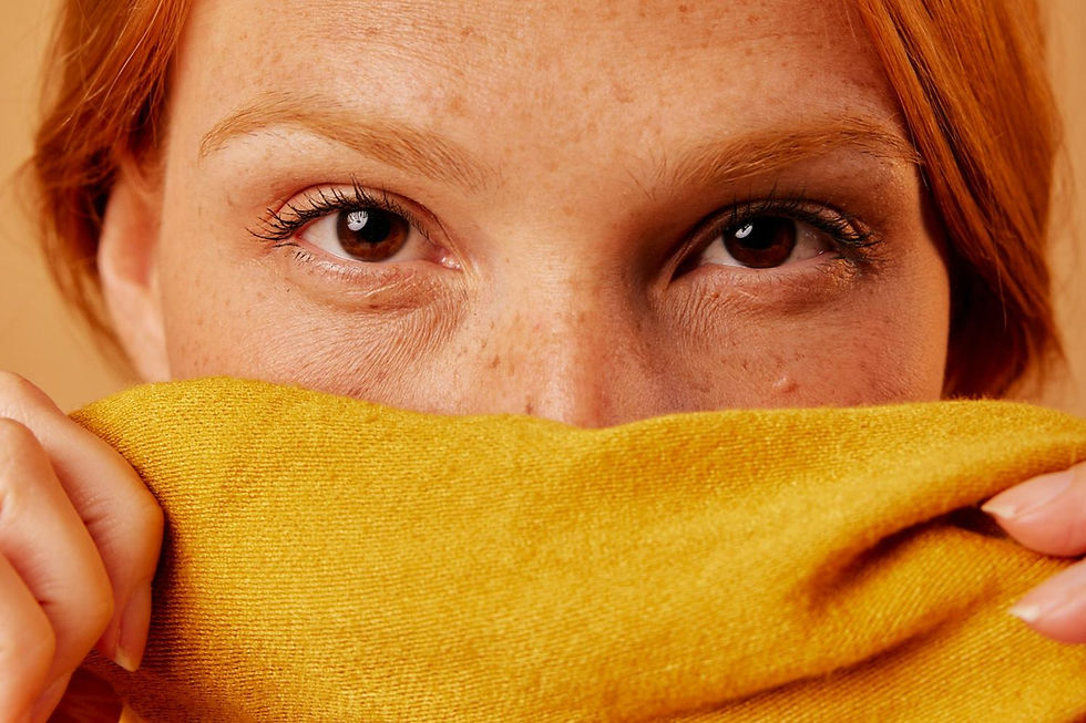 Close-up of a person with red hair partially covering their face with a yellow fabric. Brown eyes gaze forward, set against a neutral background.