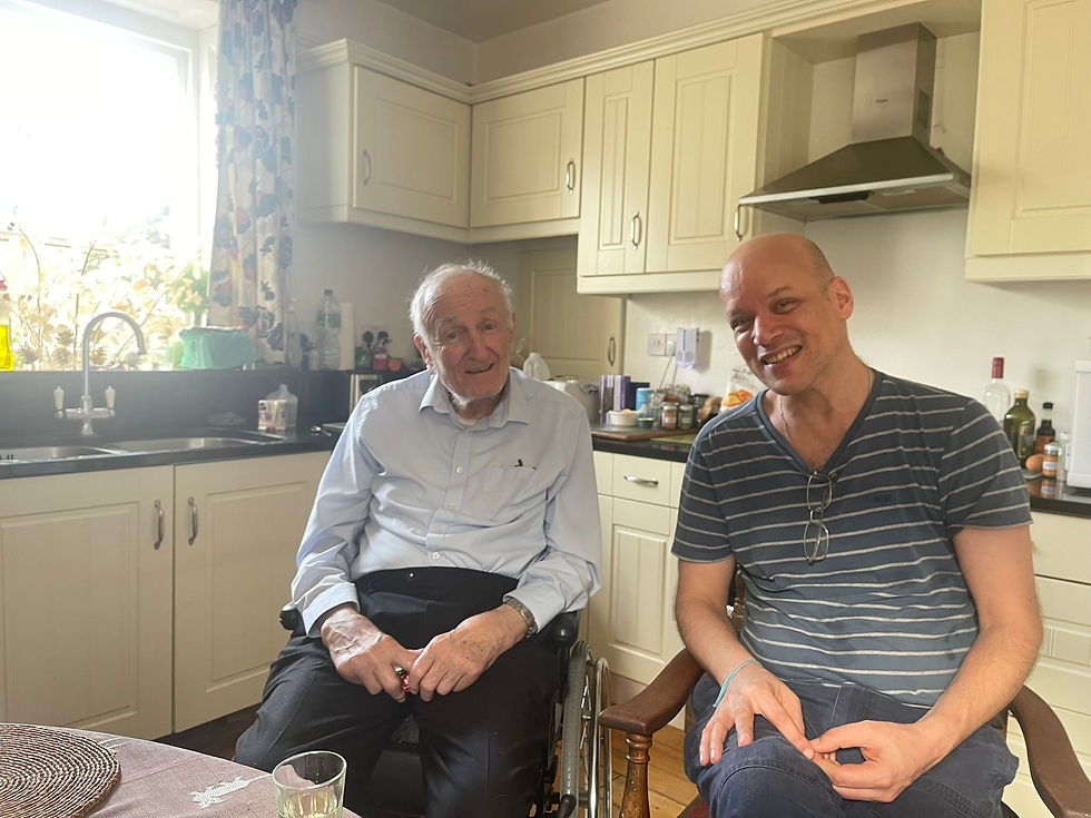 Two men smiling in a kitchen with light-colored cabinets. One is in a wheelchair. A window with floral curtains brightens the room.
