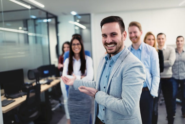 Smiling man in a suit gestures towards a line of people in an office. They stand near desks and computers, creating a welcoming atmosphere.