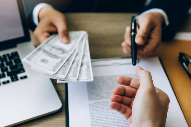 Hands exchange cash and a pen over a document on a desk with a laptop, suggesting a business transaction.