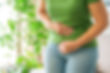 Person in green shirt and jeans clutching abdomen in discomfort. Indoor setting with green plants in background, conveying pain or upset mood.