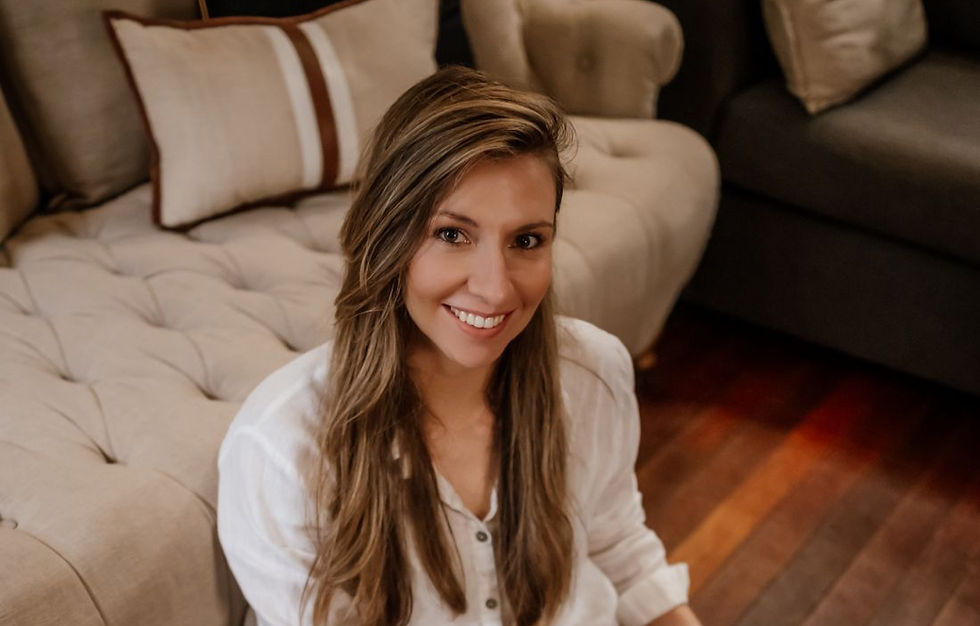 A woman with long brown hair smiles at the camera while sitting on a beige couch.