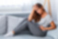 Woman in gray loungewear sits on a couch, holding her stomach with one hand on her head, appearing distressed. Bright, airy room.