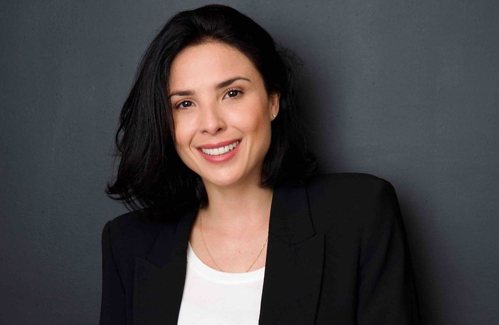 Smiling person with dark hair in a black blazer and white shirt, set against a plain dark gray background. Mood is friendly and professional.