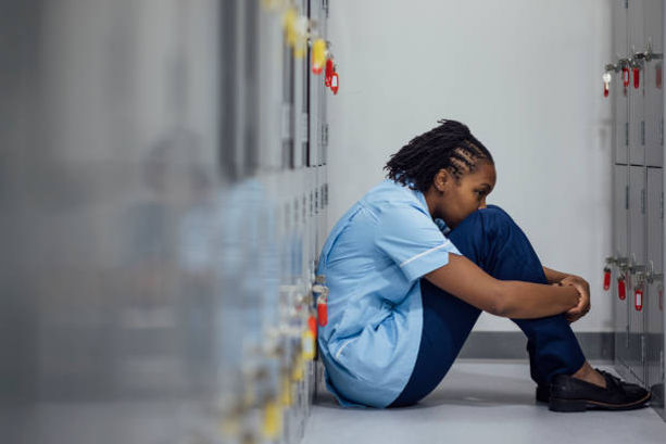The image shows a healthcare worker in blue scrubs sitting on the floor of a locker room, leaning against the lockers with her knees drawn up.