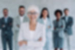 Smiling elderly woman in glasses and gray suit stands confidently, surrounded by diverse colleagues in similar attire, arms crossed.