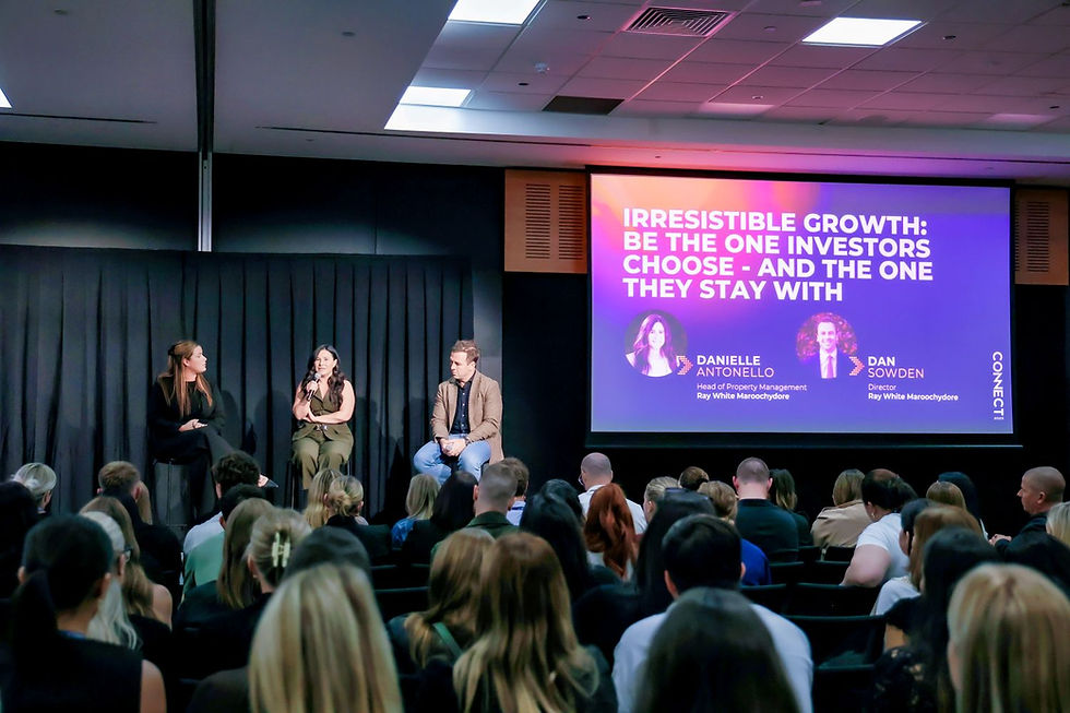 A panel of speakers presents at a business conference, addressing an audience, with a screen behind them displaying the session title and speaker names.