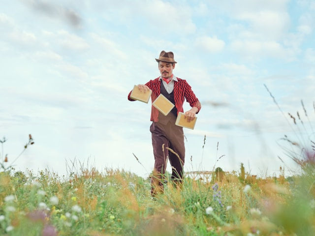 A man in vintage attire juggles three books in a grassy field under a bright sky.