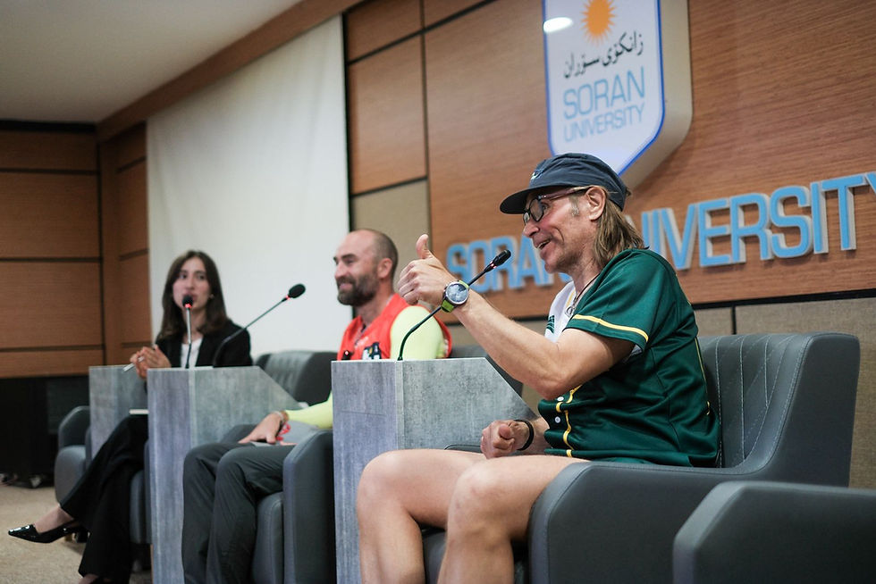 Three panelists at Soran University sit at microphones; a man in green gives a thumbs-up during a lively discussion.