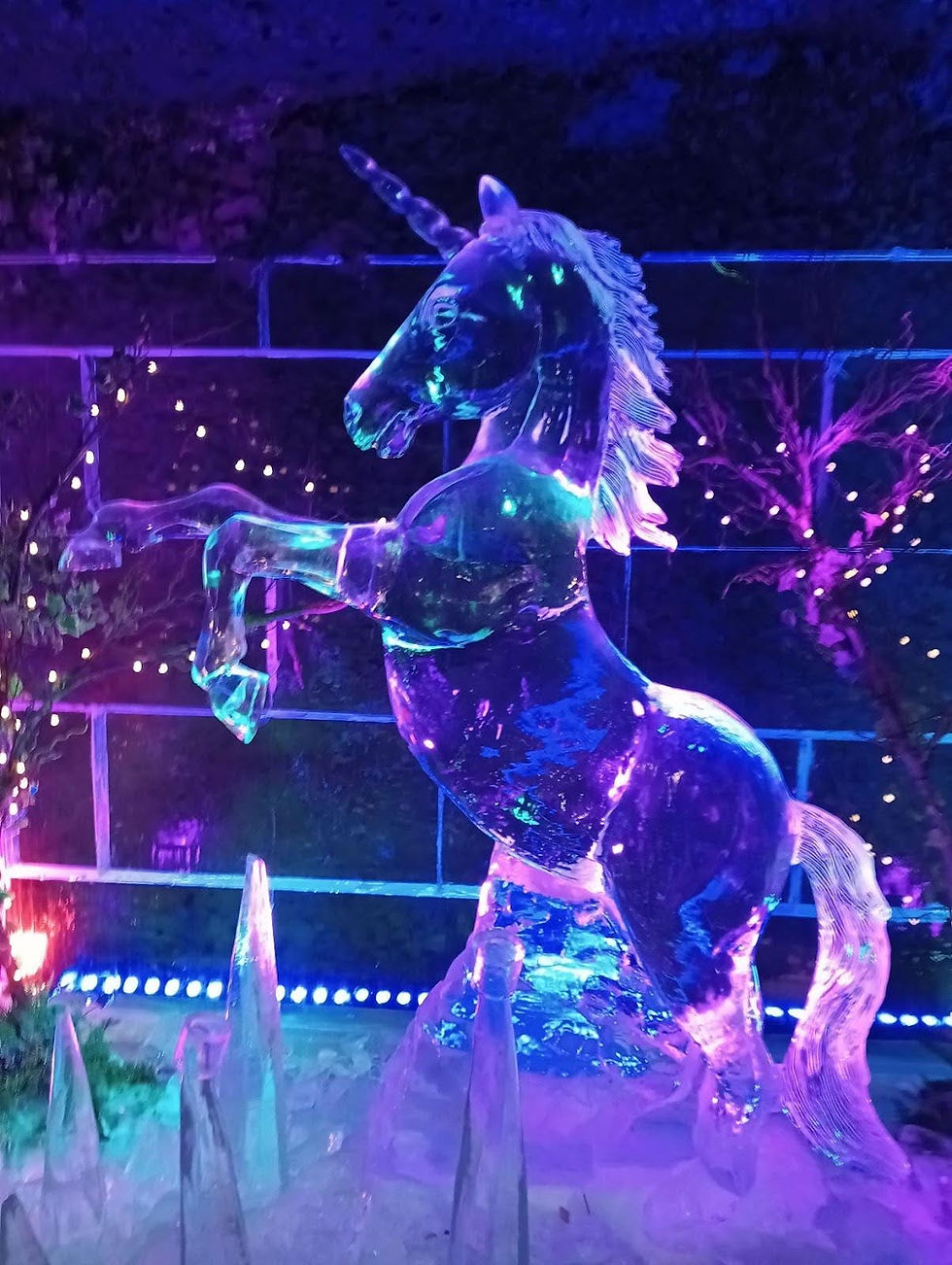 Ice sculpture of a unicorn rearing up, illuminated by purple and blue lights. Twinkling lights and icicles create a magical ambiance.
