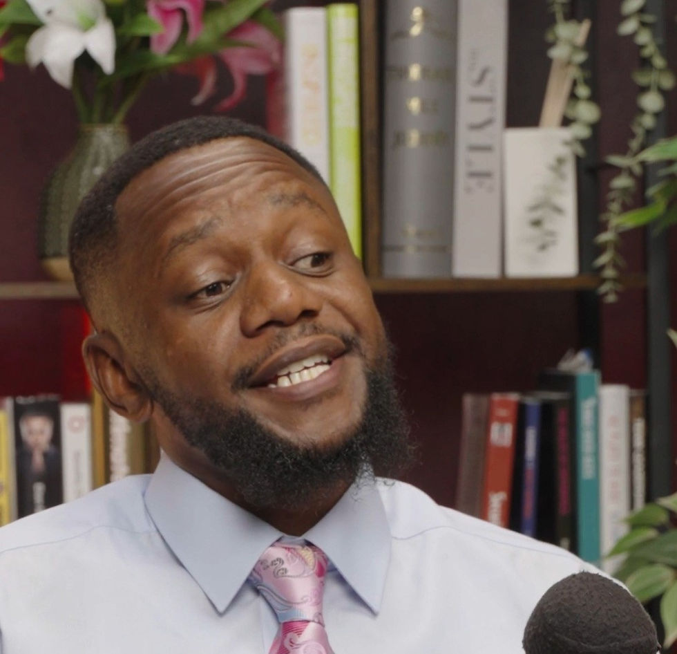 Man in a shirt and pink tie speaking into a microphone, in front of a bookshelf with colorful books and a vase of flowers.