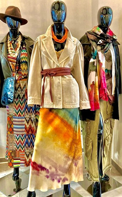 Three mannequins display layered bohemian outfits in a boutique, with hats, scarves, and tie-dye fabrics.