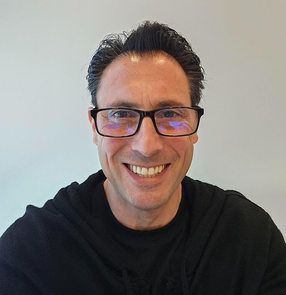 Man with glasses smiling against a plain background. He wears a black shirt. The mood is cheerful and friendly.
