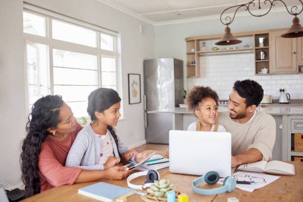 Family of four at a table in a bright kitchen, talking and smiling. A laptop, headphones, and notebooks are on the table. Warm, cozy setting.