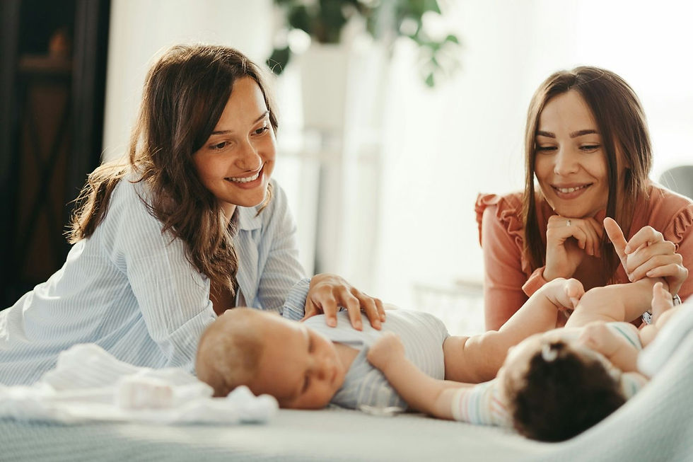 Two women smiling at two babies lying on a bed in a bright room. The scene conveys warmth and happiness.