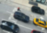 A person on a tiny motorbike rides in traffic alongside cars and a taxi on a city street.