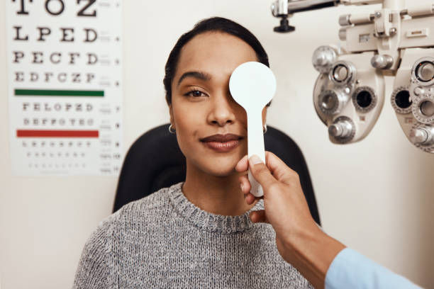 Woman in a gray sweater at an eye exam with one eye covered. Eye chart and test machine visible. Neutral expression.