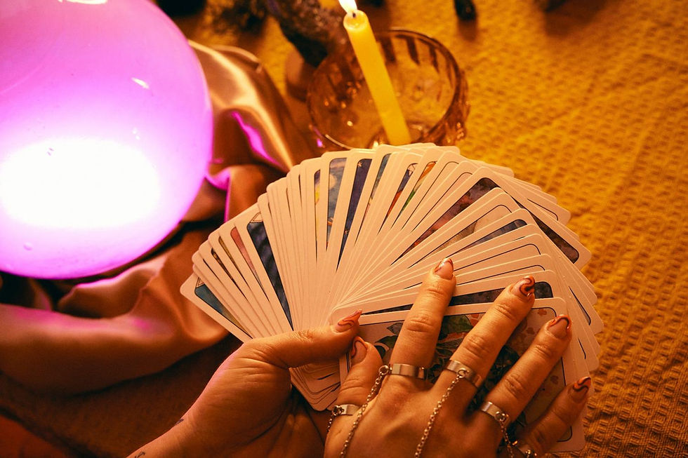 Hands with rings hold spread tarot cards near a glowing pink sphere and lit candle on a yellow textured tablecloth, creating a mystical mood.