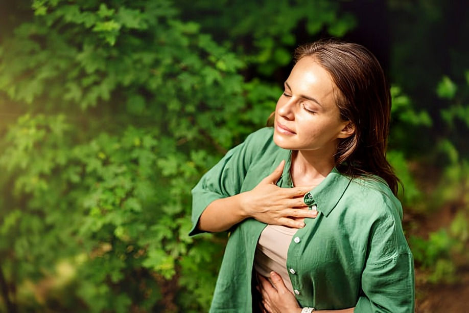 Woman in a green shirt stands outdoors among leafy trees, eyes closed and hand on chest, looking calm and reflective.
