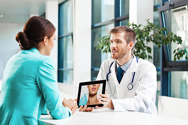 Doctor showing a tablet with a close-up image of a patient's face to a woman in a blue blazer. They're in a modern, bright office.