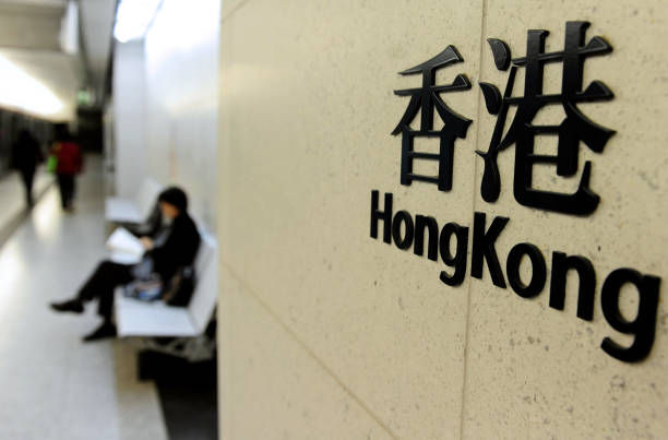 Hong Kong sign on a wall beside a blurred seated woman in a bright corridor.