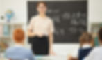 Teacher stands at chalkboard with math equations, speaking to students in a bright classroom.