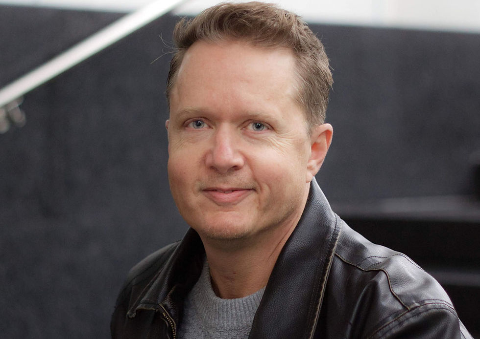Man in a leather jacket smiling slightly, sitting indoors with a blurred dark background. Casual, soft lighting, neutral mood.