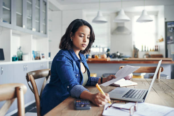 Woman studies at a kitchen table, reading papers and writing beside a laptop in a bright modern kitchen.