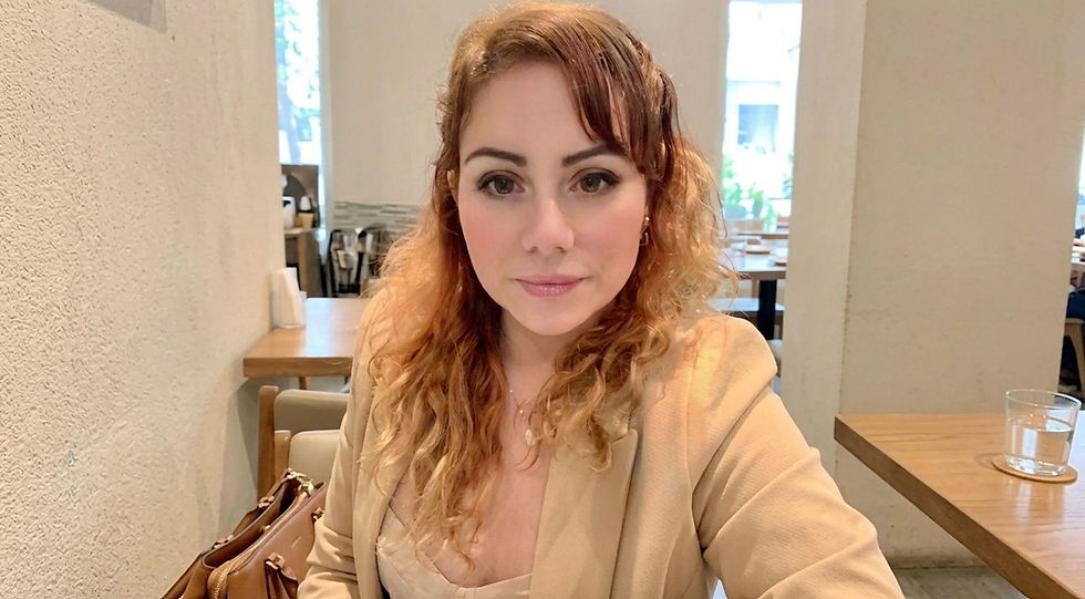 Woman in a beige blazer sits in a bright кафе, looking at the camera; tables and a glass of water in the background.