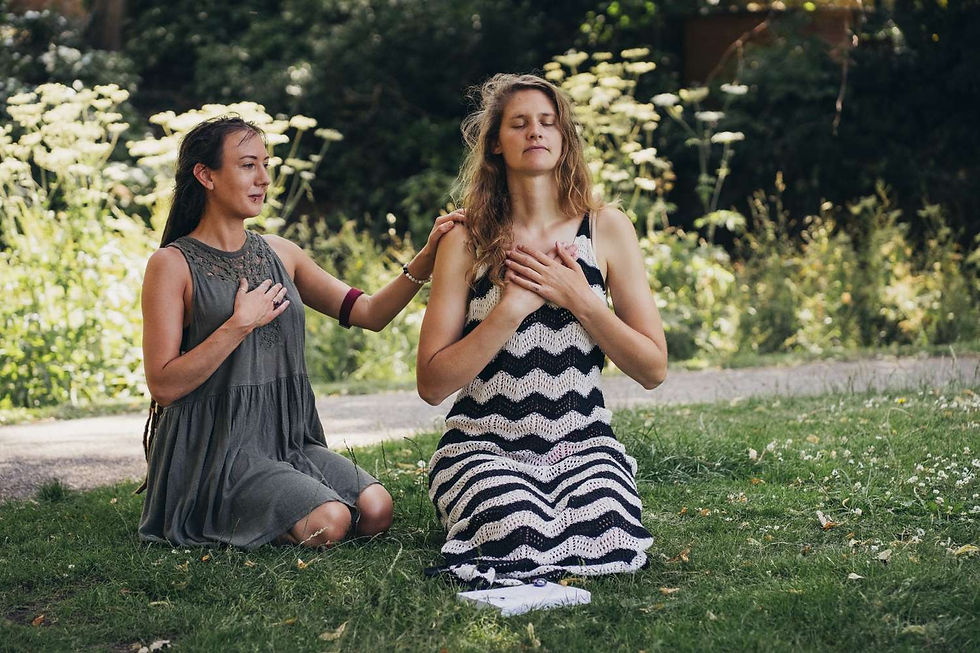 Two women kneel on grass, eyes closed, with hands on hearts. One wears a zigzag dress. Sunlit greenery in background, calm atmosphere.