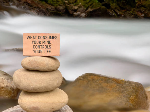 a serene stack of smooth stones by a flowing river, with a card on top reading, "What consumes your mind, controls your life."