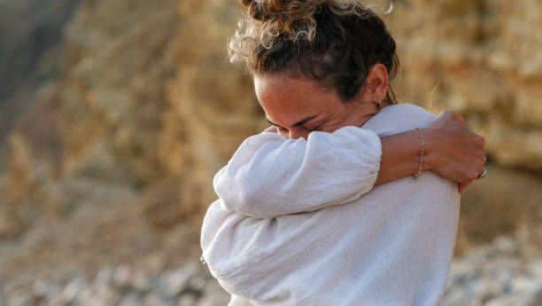 Woman in a white sweater hugs herself outdoors against a rocky background, eyes closed, conveying quiet emotion