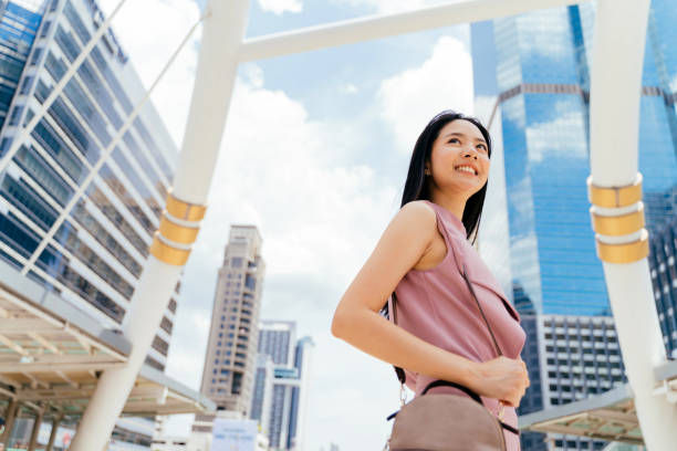 Woman in a pink top smiles, holding a bag in a cityscape. Tall glass buildings and blue sky infuse a bright, optimistic mood.